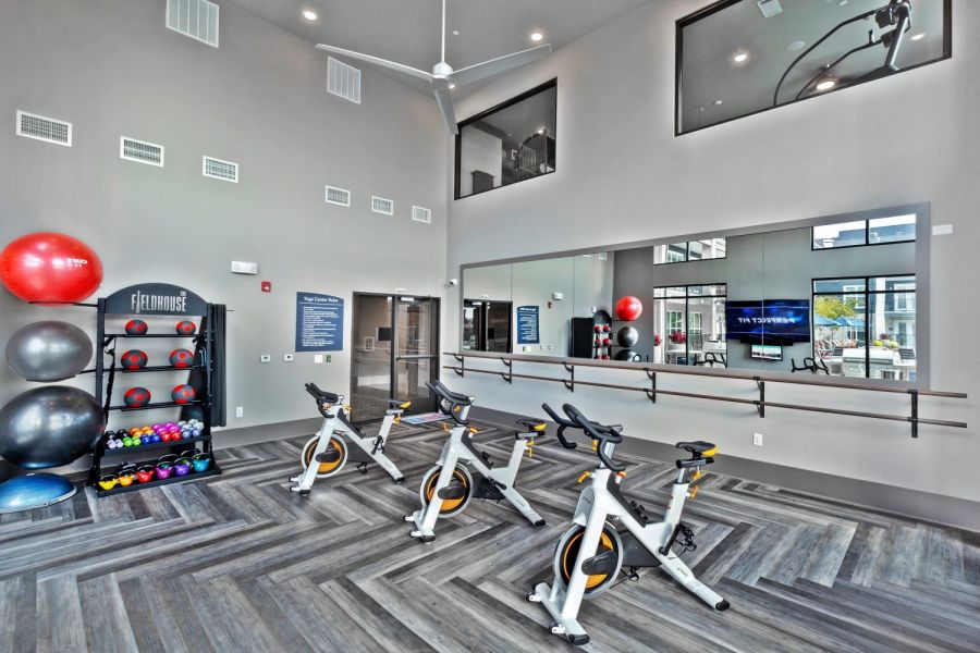 Modern fitness studio with three stationary bikes, exercise balls, weights, large mirrors, and wood-patterned flooring at The Fieldhouse Apartments.