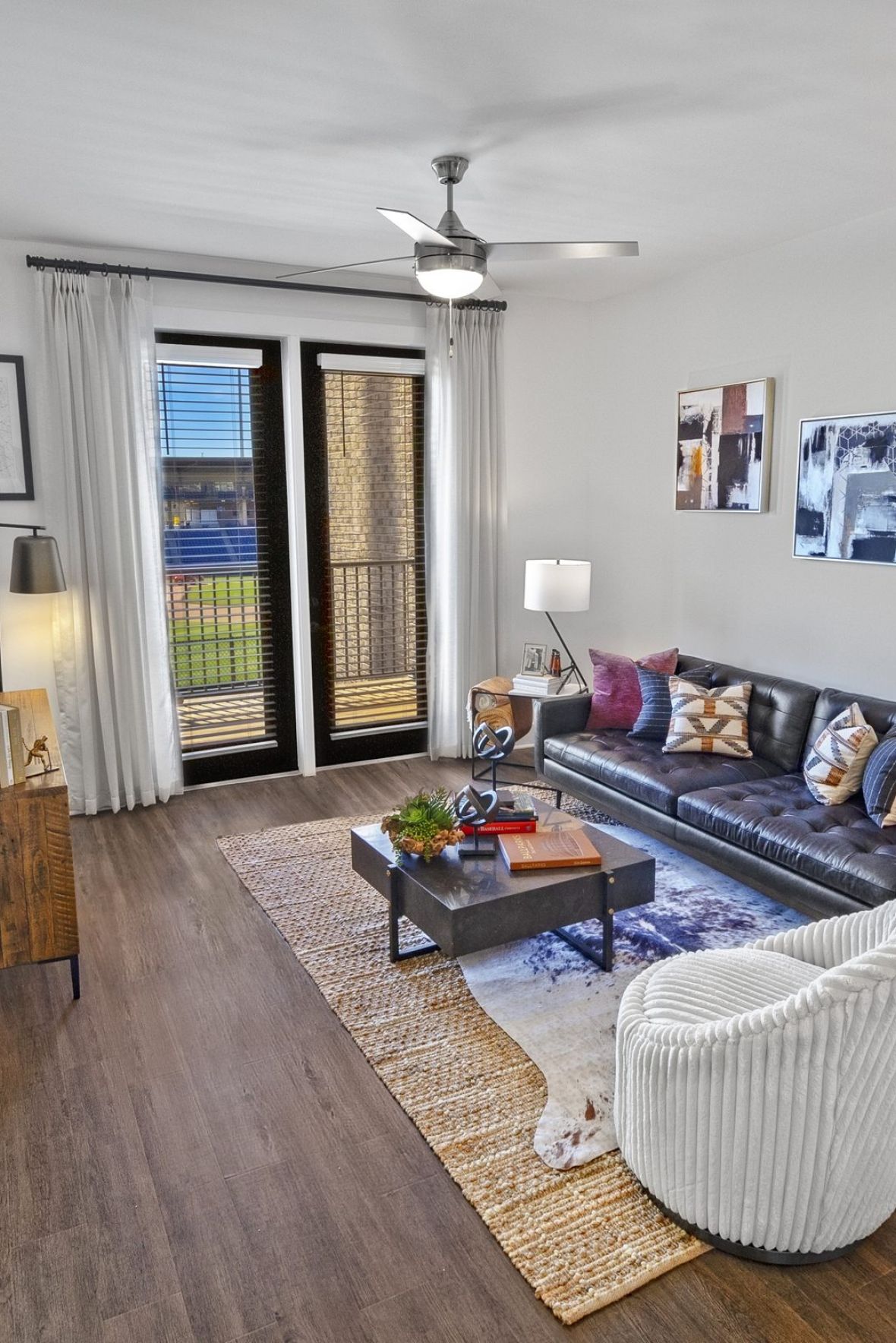 Modern living room with a gray sofa, armchair, coffee table, and large windows letting in natural light at The Fieldhouse Apartments.