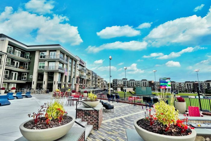 Modern apartment complex with outdoor seating overlooks a baseball field under a bright blue sky with clouds.