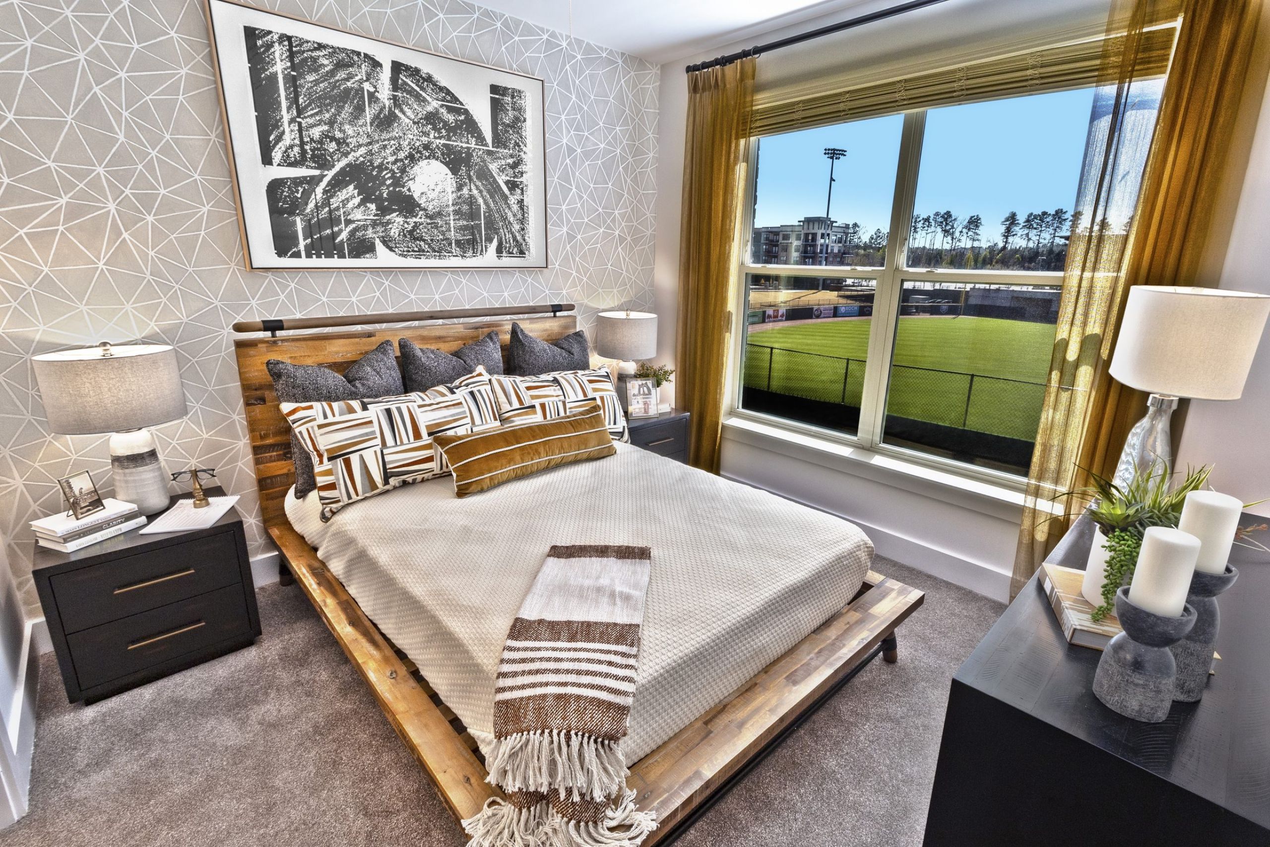 Modern bedroom with geometric wallpaper, cozy bed, lamps, and a window overlooking a sports field at The Fieldhouse Apartments.