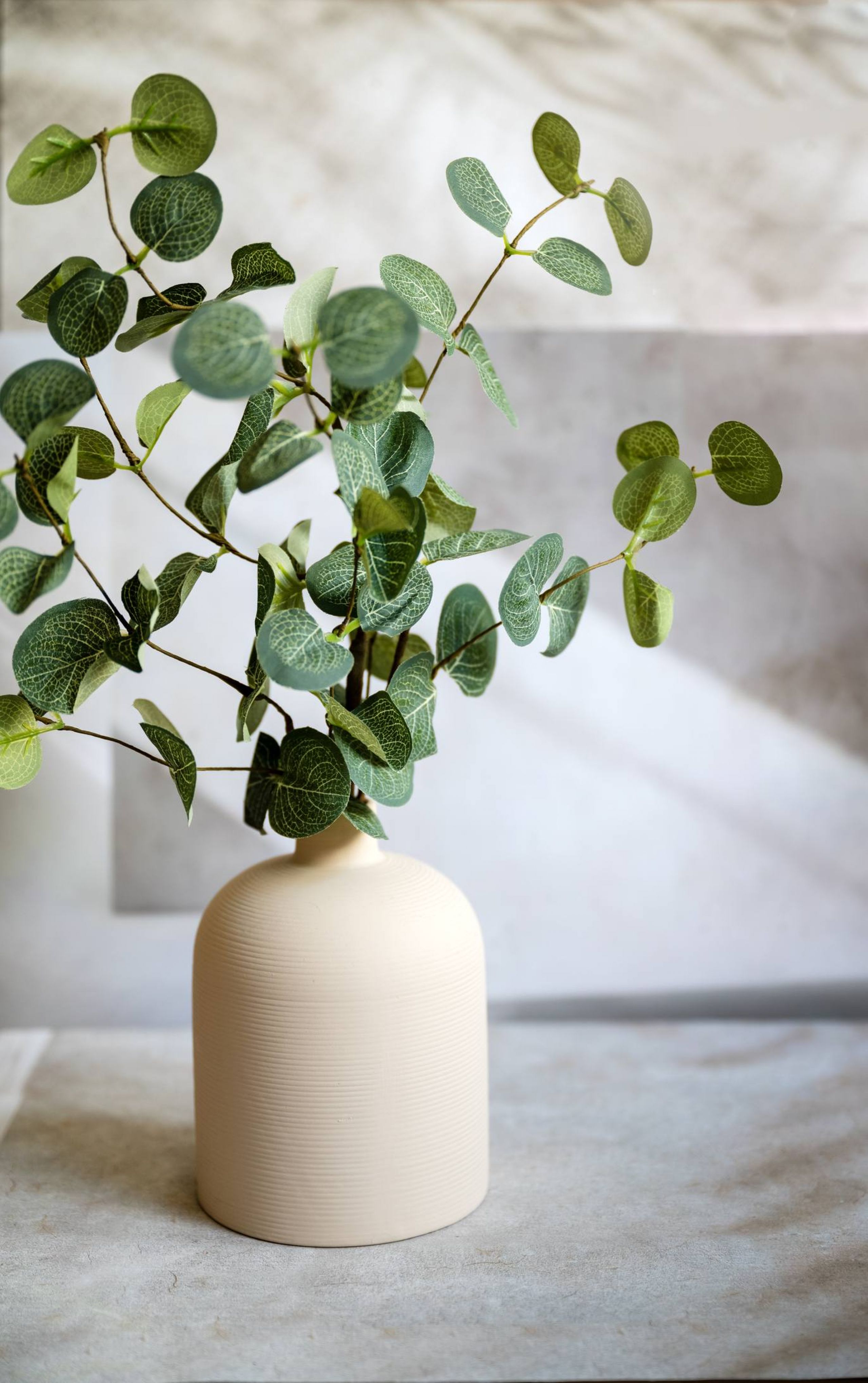 A beige vase with green leafy branches on a light-colored surface against a soft, neutral background.