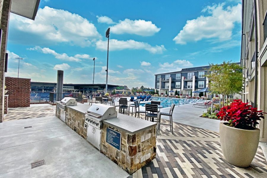 Outdoor apartment pool area with BBQ grills, tables, chairs, and modern buildings under a blue sky at The Fieldhouse Apartments.