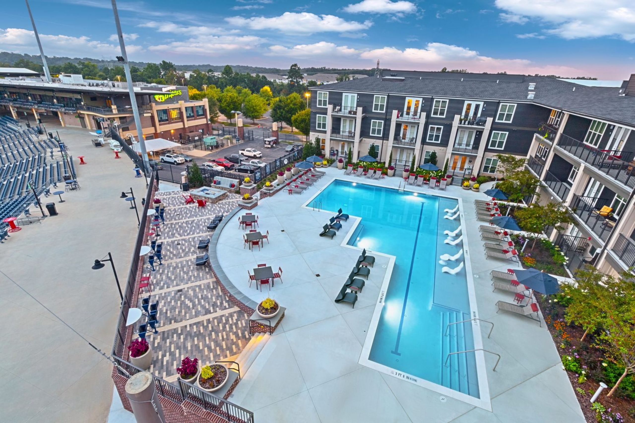 A large outdoor pool with lounge chairs beside an apartment building and a stadium under a blue sky.