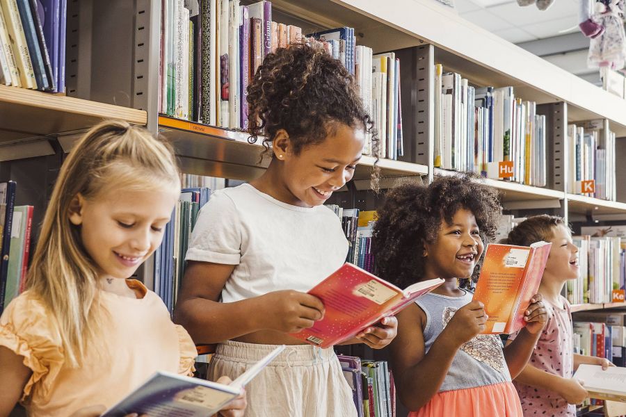 Four children standing in a library, smiling and reading books in front of shelves filled with books.