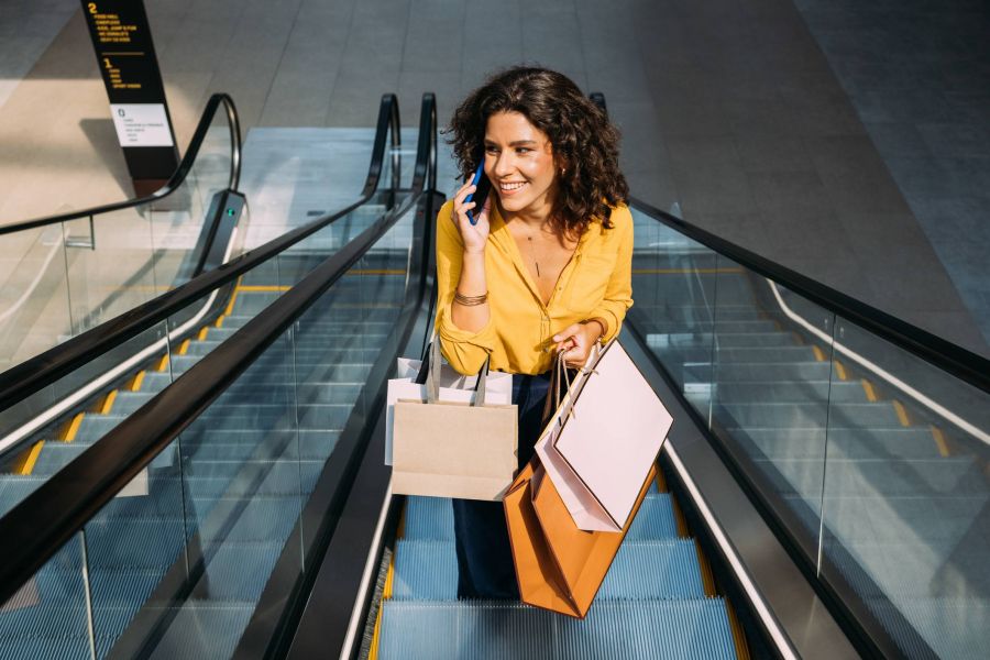 Woman with curly hair on escalator, smiling, talking on phone, and holding several shopping bags.
