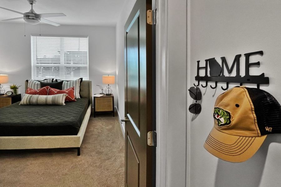Open door reveals a cozy bedroom with a bed, lamps, and a wall hook holding a hat and sunglasses at The Fieldhouse Apartments.
