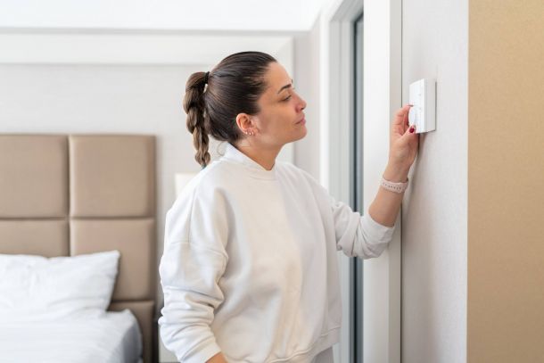 Woman in a white sweater adjusts a thermostat on a wall in a bedroom.