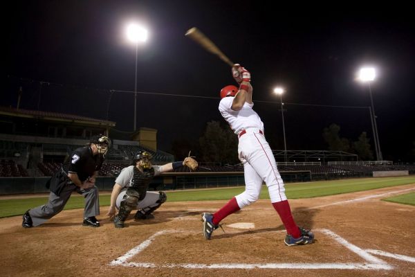 Baseball player in red uniform swings bat at night game, with catcher and umpire behind home plate.