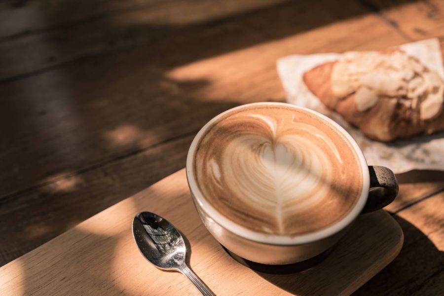 A cup of latte with heart-shaped foam art and a spoon beside it, with a croissant in the background.