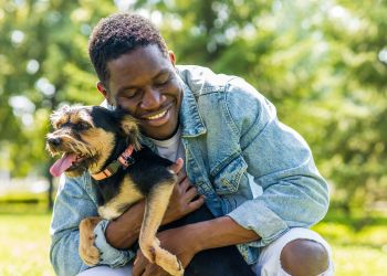 Smiling man in a denim jacket hugs a happy dog outdoors on a sunny day.