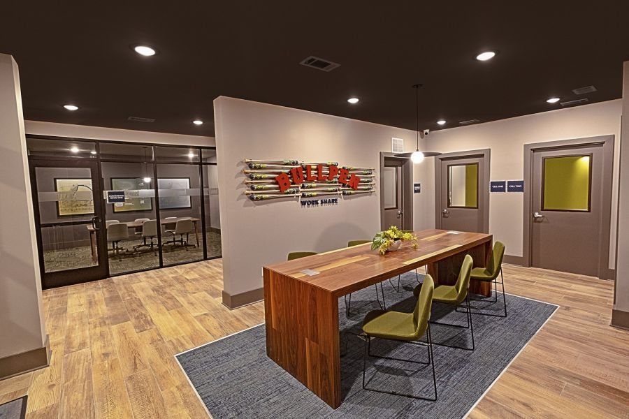 Modern office space with a wooden table, green chairs, wall art, and glass-walled meeting room in the back at The Fieldhouse Apartments.