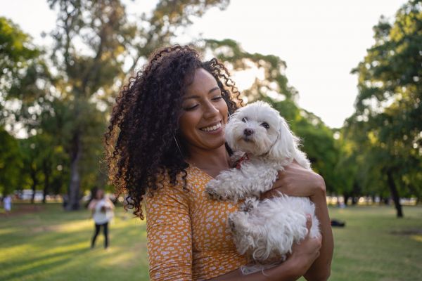 Smiling woman in a yellow dress holding a fluffy white dog at a park on a sunny day.