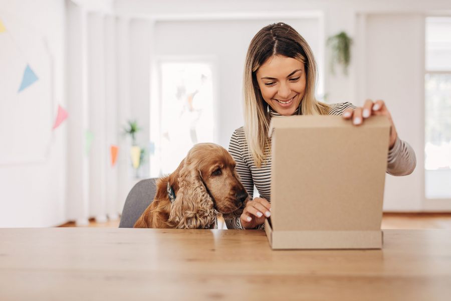 Smiling woman and her dog opening a cardboard box together at a table in a bright room.