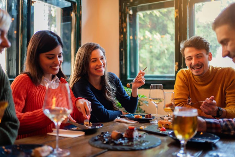 Group of friends laughing and eating together at a cozy restaurant table with drinks and food.