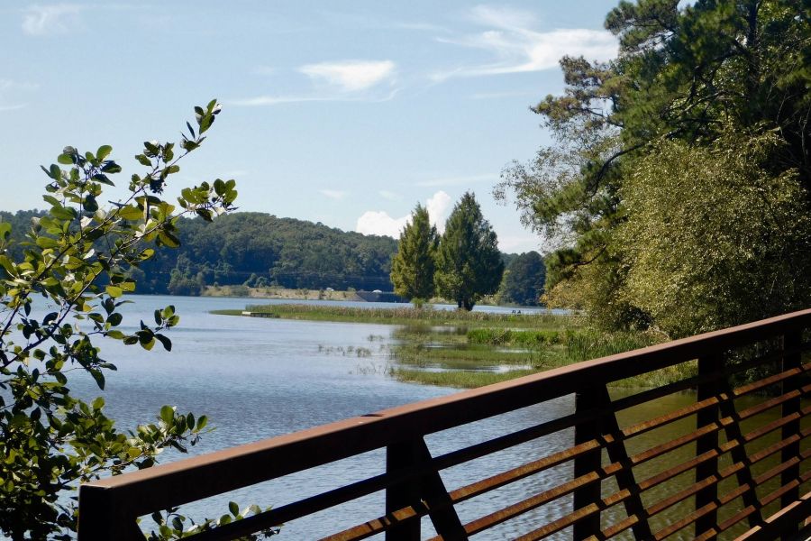 A wooden bridge crosses a lake bordered by trees under a blue sky with scattered clouds.