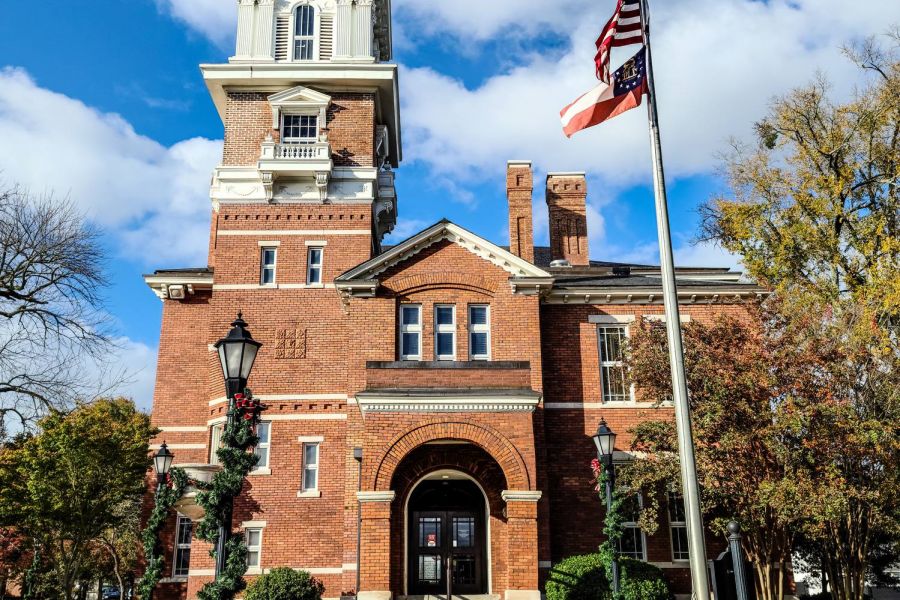 Red brick courthouse with a clock tower and two flags, under a blue sky with clouds at The Gwinnett Historic Courthouse in Lawrenceville, West Crogan Street in Gwinnett County, Georgia.