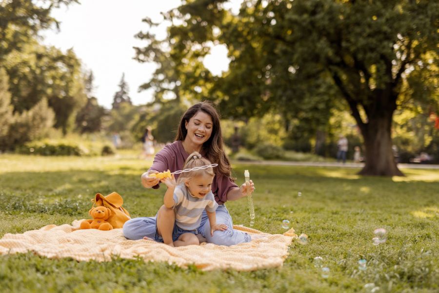 Woman and toddler sitting on a blanket in a park, smiling and playing with bubbles on a sunny day.