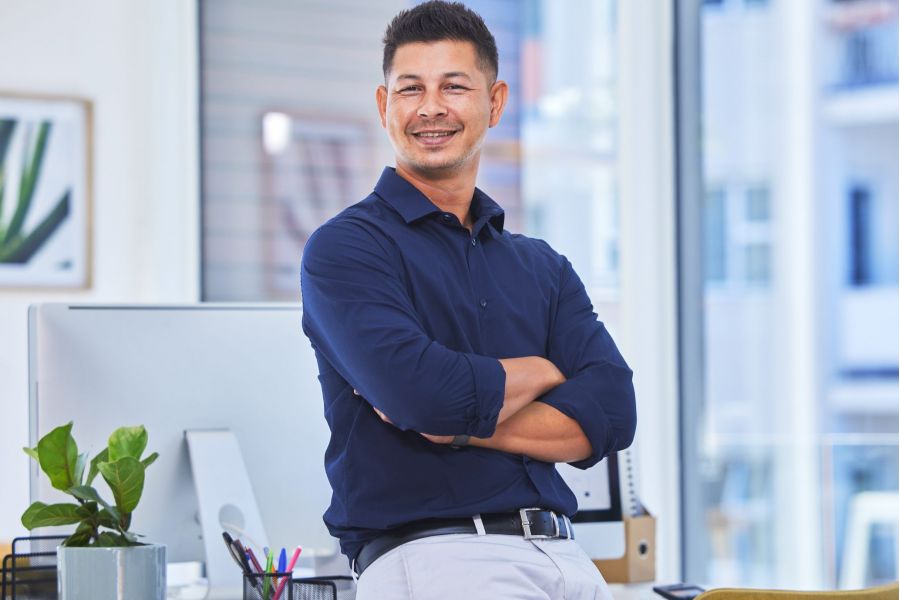 Man in a navy shirt smiling with arms crossed, standing in a modern office by a desk and computer.