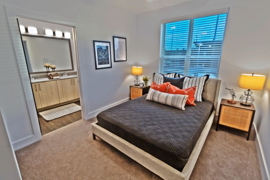 Modern bedroom with a dark bedspread, orange pillows, two lamps, and a view into a bathroom  at The Fieldhouse Apartments.