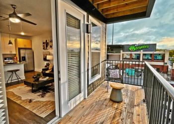 Modern apartment balcony with wooden floor, chair, and view into cozy living room and street with grocery store sign at The Fieldhouse Apartments.